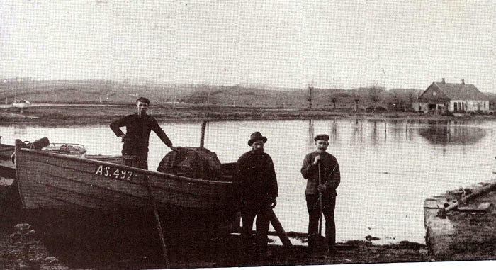 Norsminde havn omkring 1920. Her hørte nogle få fiskerbåde hjemme. Båden, der her er trukket på land, ejes af Emil Olsen, som står i båden. De to andre er ukendte. I baggrunden ses "Fisker Mines hus" (Vilhelmine Pedersen).