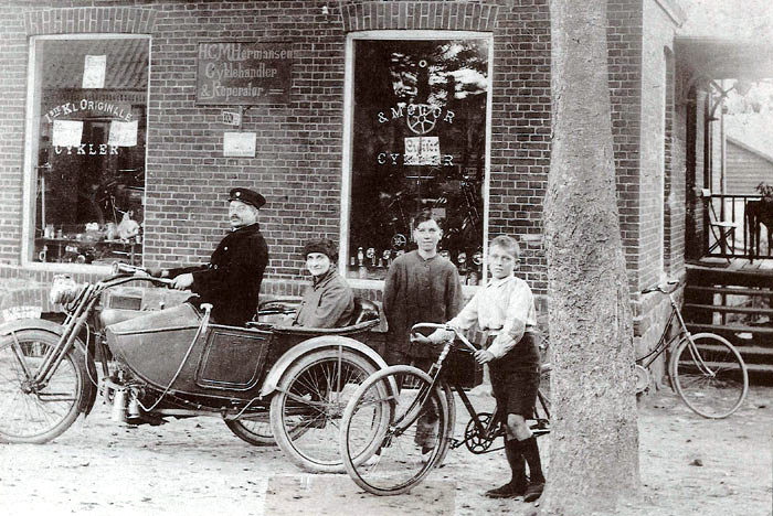 Cykelhandler Hans Christian Hermansen med familien uden for forretningen, Bredgade 66 i Malling. H. C. Hermansen sidder på motorcyklen med sin kone i sidevognen. Stående er det sønnerne Harald og Henry med cyklen. Billedet er fra omkring 1915. 