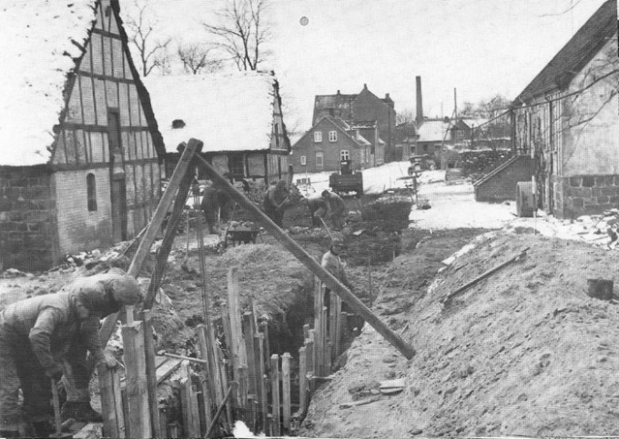 Opgravning af kloak til Malling skole, der blev taget i brug august 1959. Billedet her er fra d. 14. dec. 1957, og med den seneste kloakering i erindring må dette kaldes primitivt, men det fungerede. Gennembrydningen foregår ved "Lundshøj gård", hvor stuehuset til højre nu er præstebolig, mens der er P-plads, hvor udbygningerne lå. Vejen er nu forbindelsesvej mellem Kirkevej og Lundshøjgårdsvej.